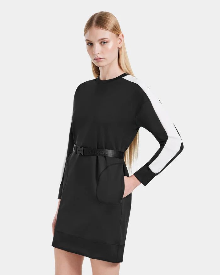 A woman with long, straight blonde hair models the Rudsak SETA - 8525019 Black mini dress, featuring white sleeve stripes and a belted waist, standing with one hand in her pocket against a plain white background.