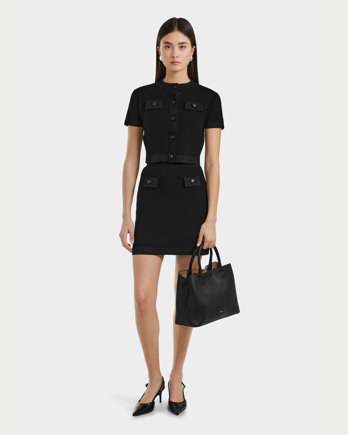 A woman in a short-sleeve black buttoned dress with front pockets, black pointed heels, and a Rudsak SIDNEY - 8525074 Black sweater draped over her shoulders stands against a plain white background.