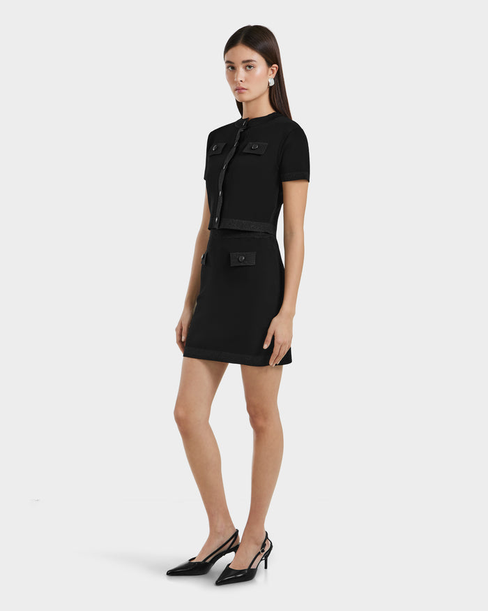 A woman with straight brown hair and a neutral expression wears the Rudsak SIDNEY - 8525074 Black metallic-trim sweater over a short-sleeved black dress with front pockets and buttons, paired with black pointed-toe heels, against a plain white background.