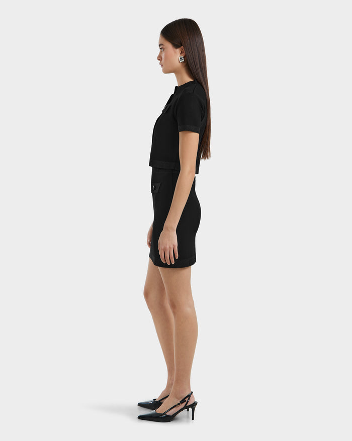 A woman in profile faces left, wearing the Rudsak SIDNEY - 8525074 Black fitted cropped sweater and matching mini skirt, with black slingback heels and statement earrings. She has long, straight brown hair against a plain white background.