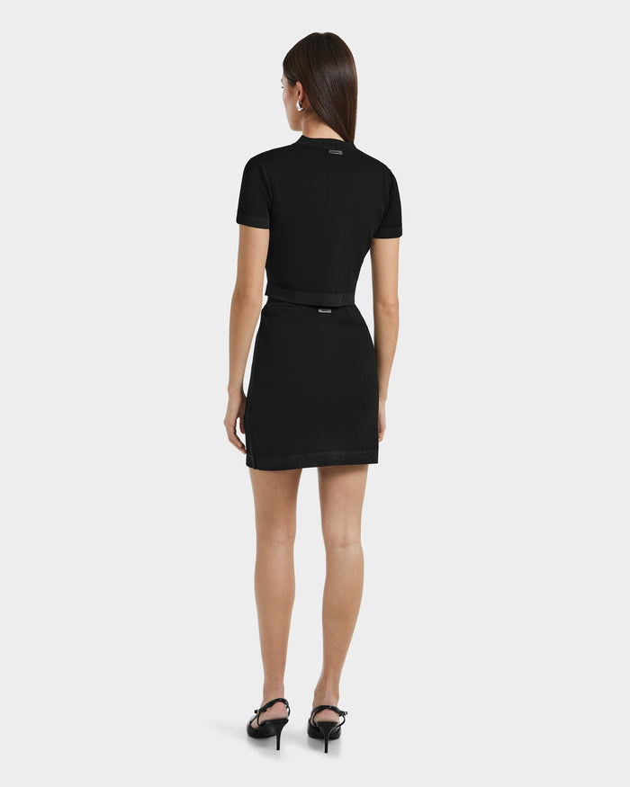 A woman with long brown hair faces away, wearing the Rudsak SIDNEY - 8525074 Black belted dress and black high-heeled sandals against a plain white background.