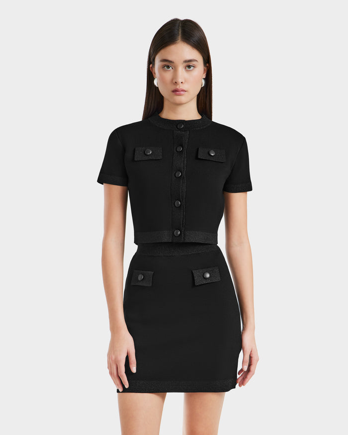 A woman wears the Rudsak SIDNEY - 8525074 Black set: a short-sleeved black button-up top with metallic trim and chest pockets, paired with a matching high-waisted mini skirt, standing against a light gray background.