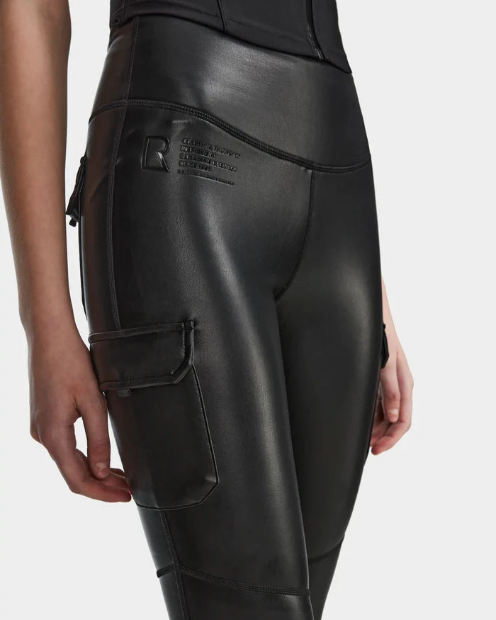 A person wears Rudsak SIEN L - 8524079 Black high-waisted, stretch vegan leather leggings with a side pocket and printed hip text, shown from mid-torso to thigh against a plain white background.