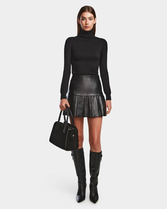 A woman faces forward in a black turtleneck, the Rudsak SOLANGE - 8524543 Black women's leather skirt, knee-high black boots, and holds a black handbag against a plain white background.