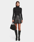 A woman faces forward in a black turtleneck, the Rudsak SOLANGE - 8524543 Black women's leather skirt, knee-high black boots, and holds a black handbag against a plain white background.