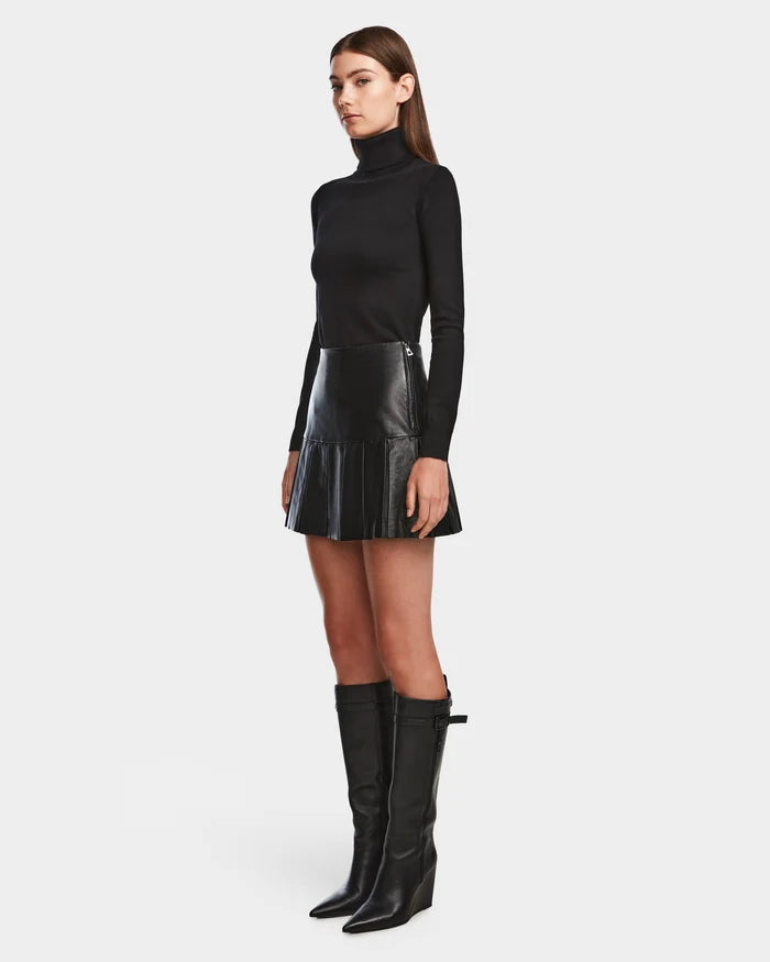 A woman stands against a plain background wearing the Rudsak SOLANGE - 8524543 Black pleated leather skirt, a black turtleneck, and knee-high black boots, facing forward with a neutral expression.