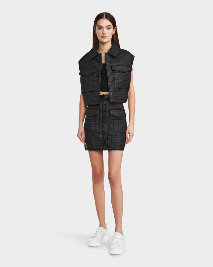 A woman stands facing forward against a white background, wearing the Rudsak SOLSA - 8124048 Black women's cropped quilted vest with eco-friendly Sorona fill, paired with a matching black skirt, black crop top, and white sneakers.