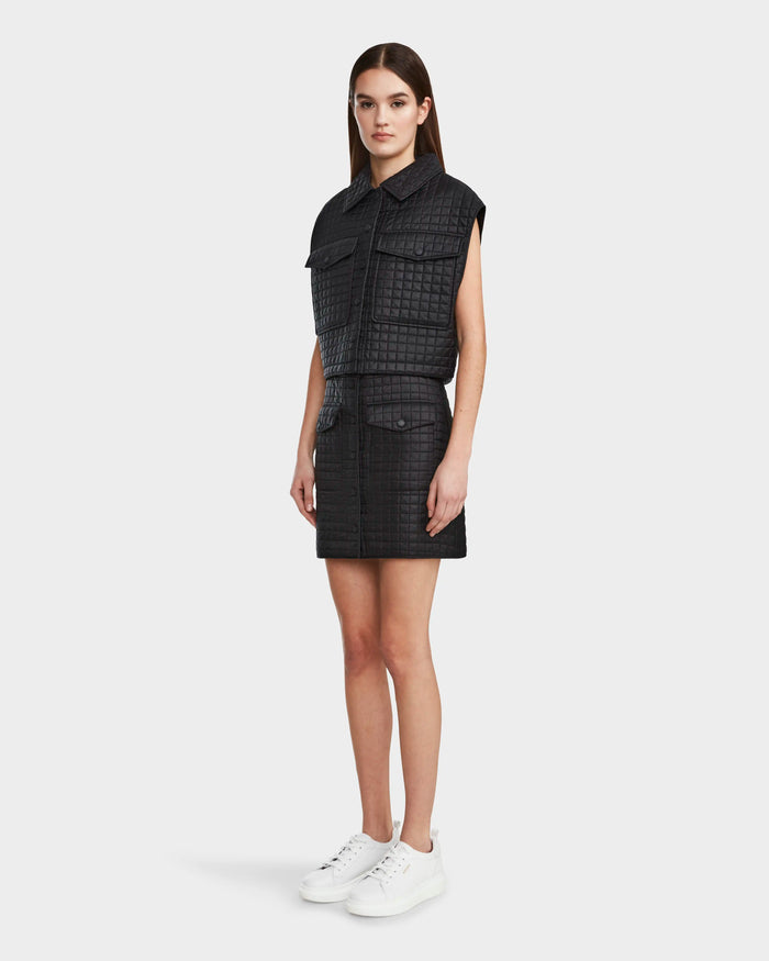 A woman poses in Rudsak’s SOLSA - 8124048 Black sleeveless, textured shirt-jacket dress with front pockets and a matching mini skirt. She wears white sneakers, has straight dark brown hair, and stands against a plain white background.