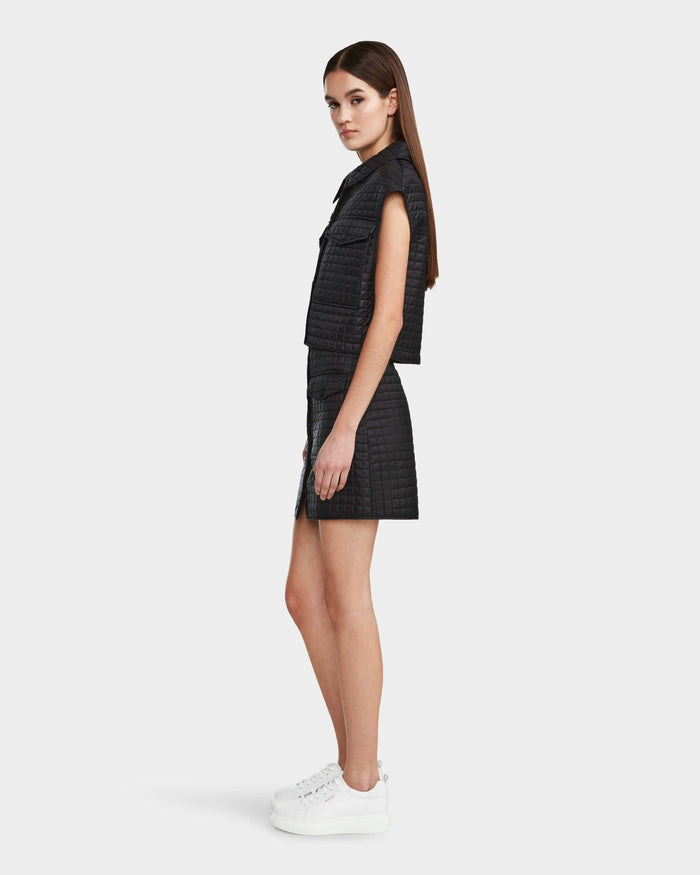 A woman with straight brown hair and a neutral expression stands in profile wearing the Rudsak SOLSA - 8124048 Black women's cropped quilted vest with eco-friendly Sorona fill, a matching skirt, and white platform sneakers against a plain white background.