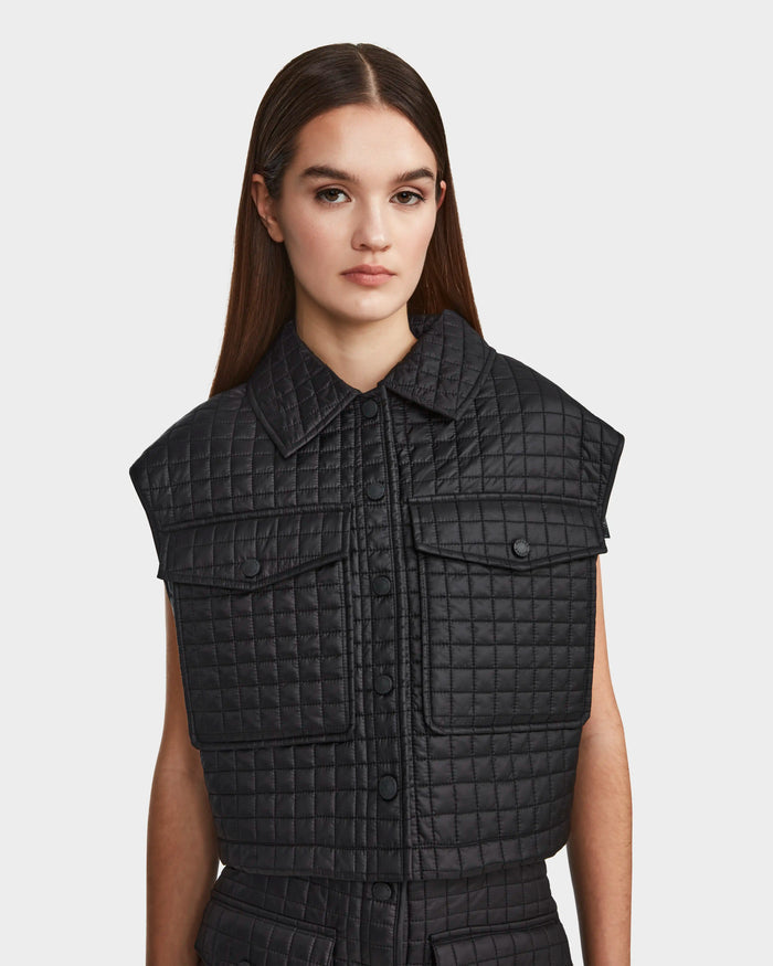 A woman with long brown hair wears the Rudsak SOLSA - 8124048 Black women's cropped quilted vest with eco-friendly Sorona fill, collar, and front pockets, standing against a plain white background.