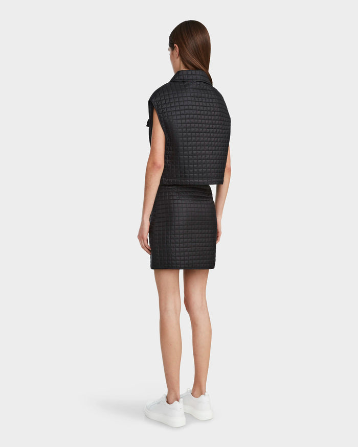 A woman with long brown hair faces away, wearing the Rudsak SOLSA - 8124048 Black textured short-sleeve jacket and matching skirt, paired with white sneakers, against a plain white background.