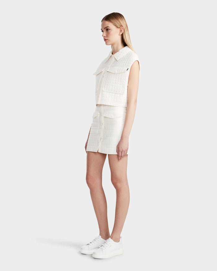 A woman in profile wears the Rudsak SOLSA - 8124048 Eggshell women's cropped quilted vest with eco-friendly Sorona fill and large front pockets, styled with a white mini skirt and sneakers. Her straight blonde hair stands out against the light gray background.