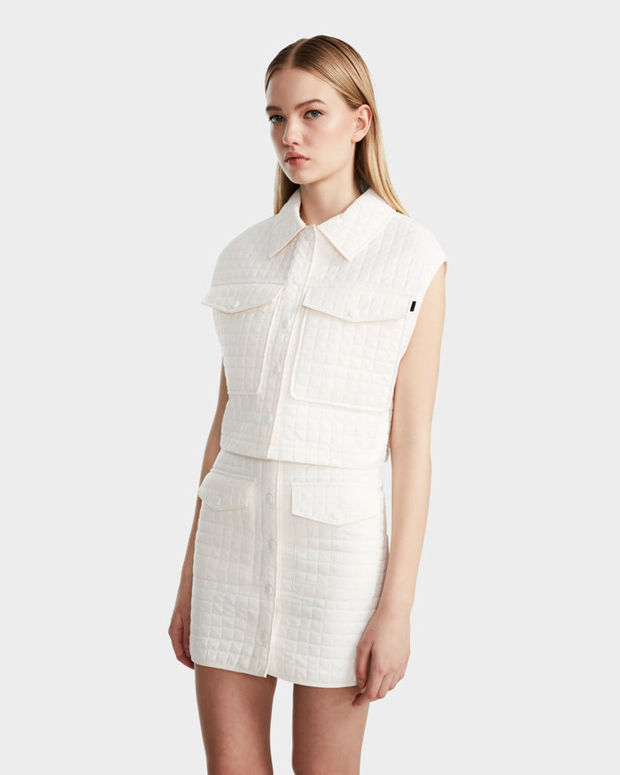 A woman with long blonde hair wears Rudsak’s SOLSA - 8124048 Eggshell, a sleeveless white cropped quilted vest with eco-friendly Sorona fill and collar, paired with a matching quilted mini skirt, standing against a plain light background.