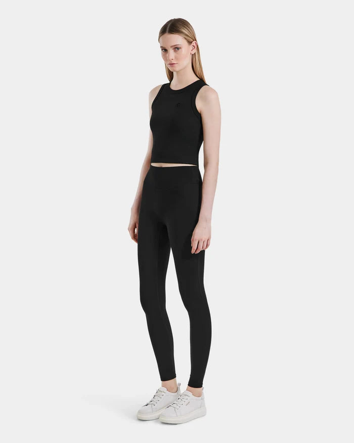 A woman with straight blonde hair and a neutral expression stands against a white backdrop, wearing the Rudsak SONOMA - 8525071 Black sleeveless crop top, workout leggings, and white sneakers, facing slightly to the side.