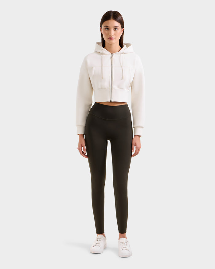 A woman faces forward against a plain light background, wearing the Rudsak SONOMA - 8525071 Dark Olive high-waisted versatile workout leggings, a white cropped zip-up hoodie, and white sneakers.