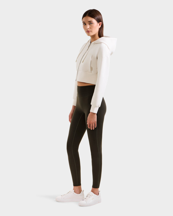 A woman stands turned slightly to the side, wearing a white cropped hoodie, Rudsak SONOMA - 8525071 Dark Olive leggings, and white sneakers against a plain light background.