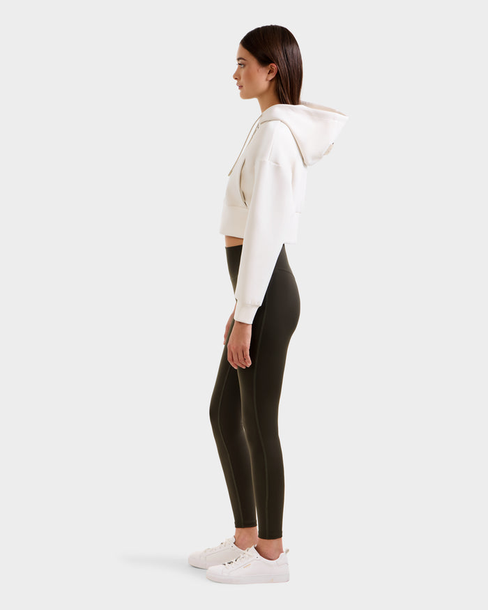 A woman stands in profile wearing a white cropped hoodie, Rudsak SONOMA - 8525071 Dark Olive leggings, and white sneakers against a plain light background.