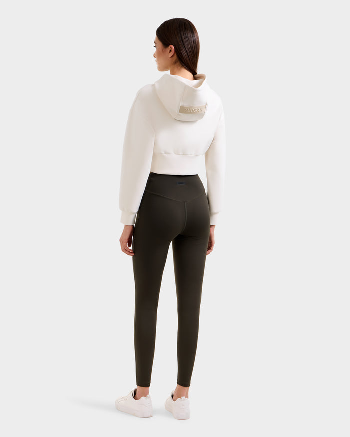 A woman with straight brown hair, seen from behind, wears a white cropped hoodie and Rudsak SONOMA - 8525071 Dark Olive leggings. She pairs the look with white sneakers against a plain light gray backdrop.