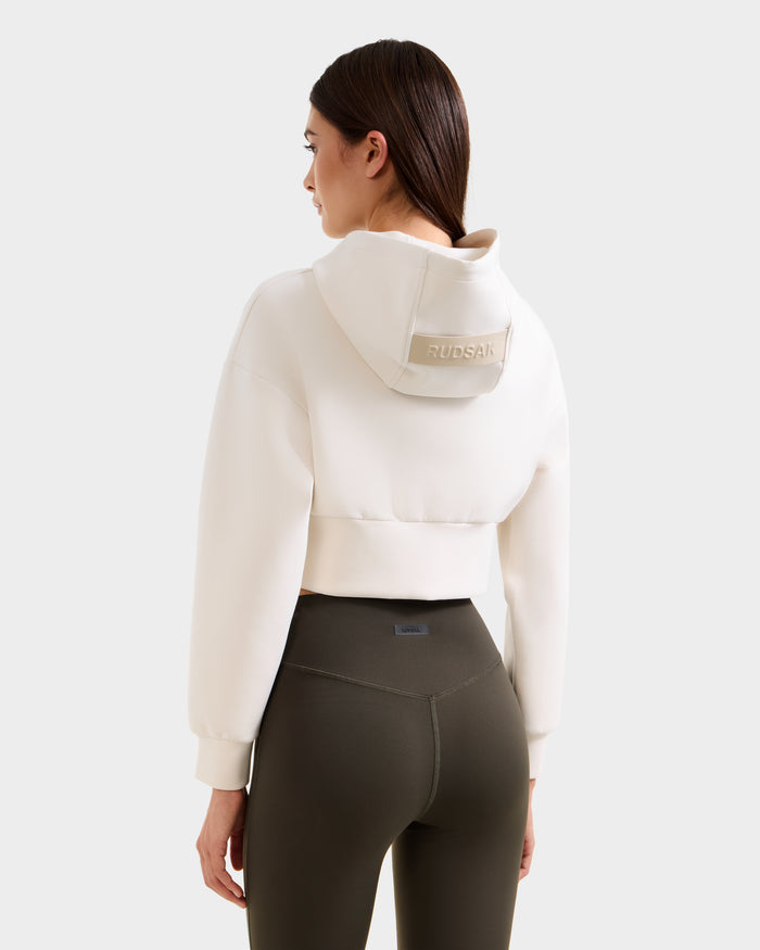 A woman faces away, wearing a cream-colored Rudsak hoodie with RUDSAK on the hood and high-waisted Rudsak SONOMA - 8525071 Dark Olive leggings, against a plain light background.