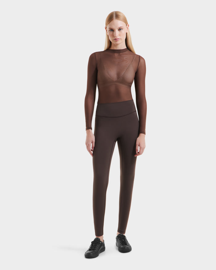 A woman with straight blonde hair and a neutral expression wears the Rudsak SONOMA - 8525071 Espresso high-waisted leggings, a sheer brown long-sleeve top over a matching bra, and black sneakers against a plain white background.