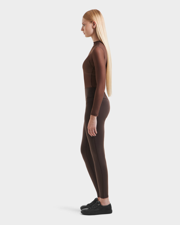 A woman with long straight blonde hair stands in profile, wearing a fitted brown top and versatile Rudsak SONOMA - 8525071 Espresso leggings, paired with black sneakers, against a plain white background.