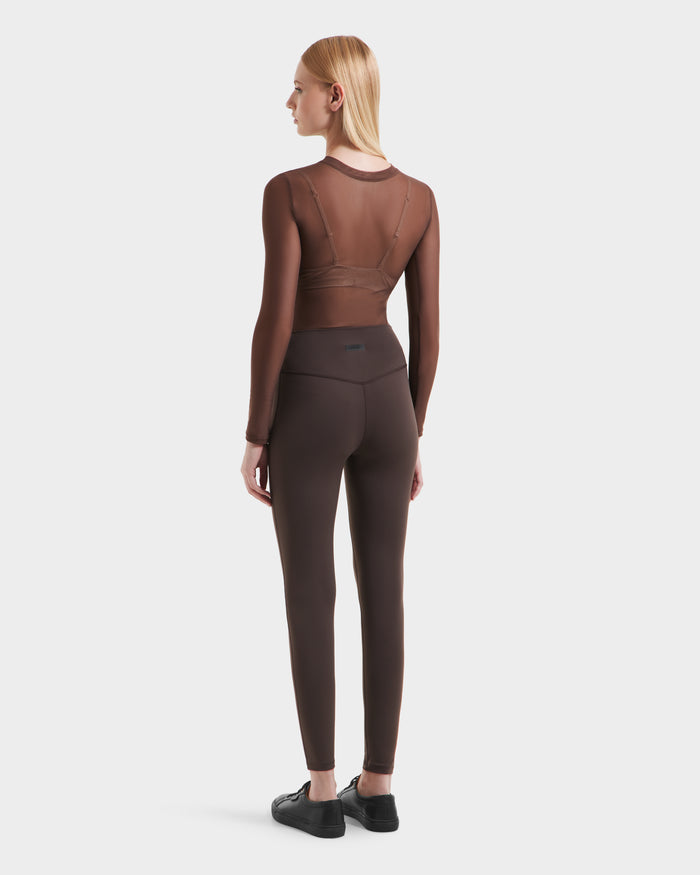 A woman with straight blonde hair, seen from behind, wears a brown mesh long-sleeve top over a matching bra and high-performance Rudsak SONOMA - 8525071 Espresso leggings with black sneakers against a plain white background.