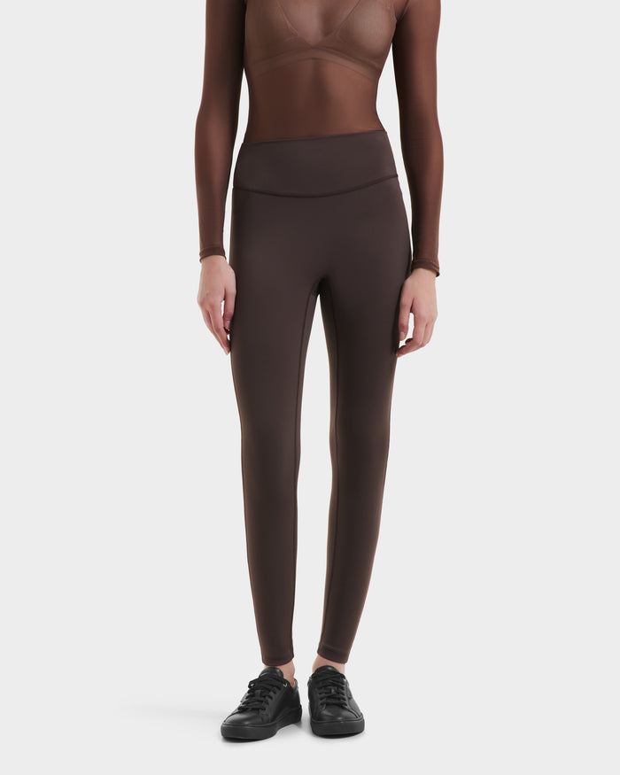 A person wearing a sheer, long-sleeved brown top and high-waisted, fitted Rudsak SONOMA - 8525071 Espresso leggings with black sneakers, standing against a plain white background.