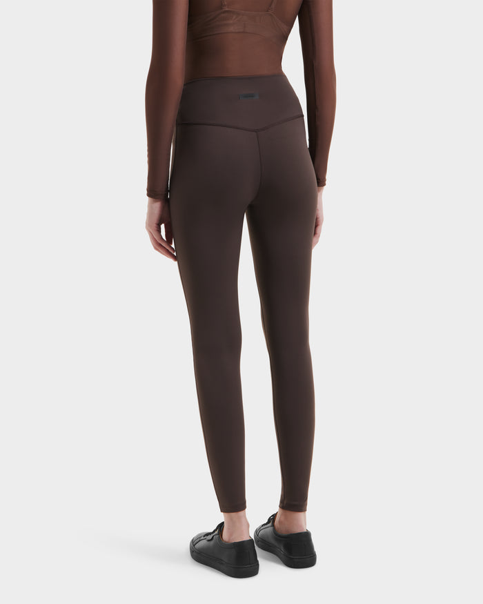A person stands facing away in a brown long-sleeve top, Rudsak SONOMA - 8525071 Espresso versatile workout leggings, and black sneakers, set against a plain white background.