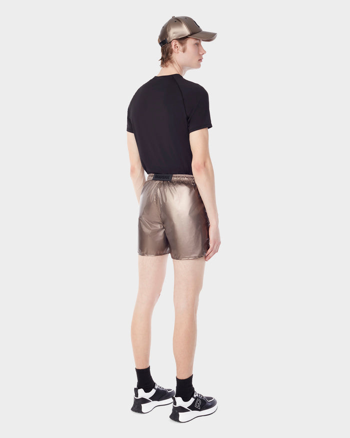 A person wearing a black T-shirt, Rudsak STANLEY - 6523006 METALLIC CHOCOLATE mens sports shorts, black socks, black and white sneakers, and a metallic cap stands facing away against a plain white background.
