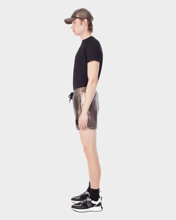 A person in profile wears a brown cap, black t-shirt, Rudsak STANLEY - 6523006 METALLIC CHOCOLATE mens sports shorts over compression shorts, black socks, and black sneakers, standing against a plain light background.