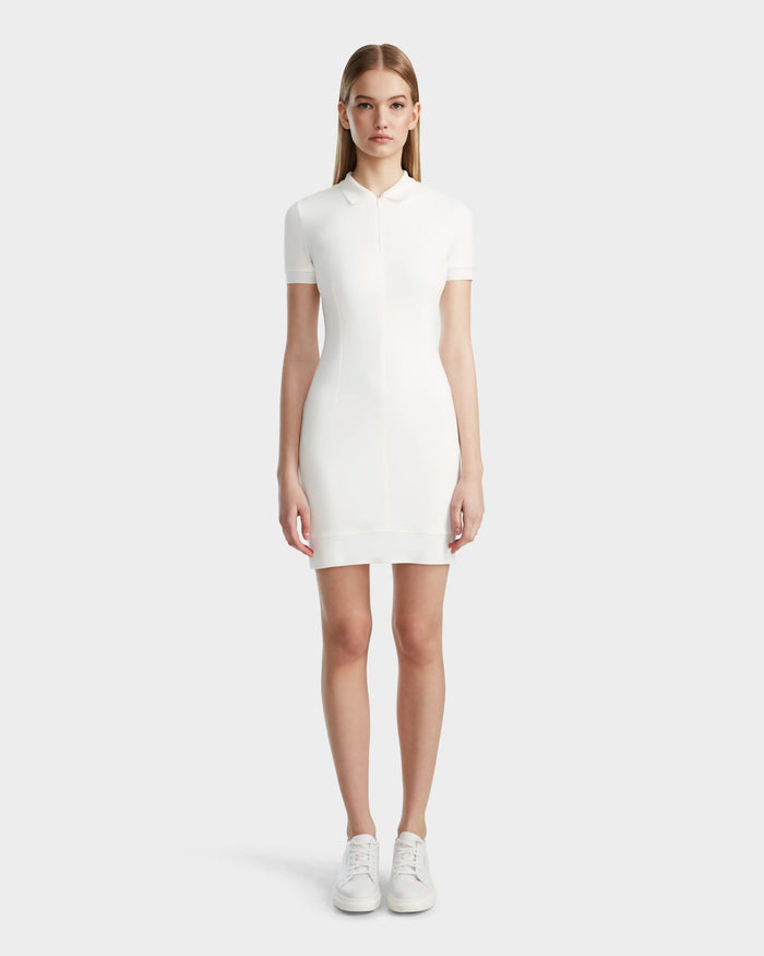 A woman faces forward wearing the Rudsak SUKI - 8524029 Cotton White fitted short-sleeve polo dress with white sneakers, showcasing sporty elegance against a plain light gray background.