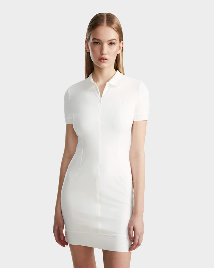 A woman with straight, light brown hair wears the Rudsak SUKI - 8524029 Cotton White women's fitted polo dress, featuring short sleeves, a collar, and zip-up front, exuding sporty elegance against a plain light gray background.