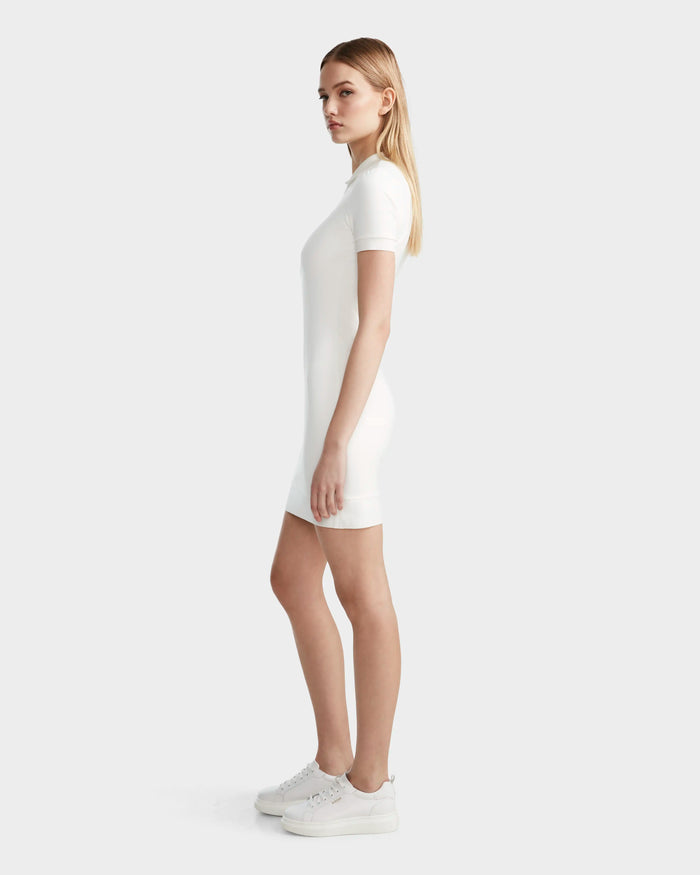 A young woman stands in profile wearing the SUKI - 8524029 Cotton White women's polo dress by Rudsak with white sneakers, radiating sporty elegance against a plain light gray background.