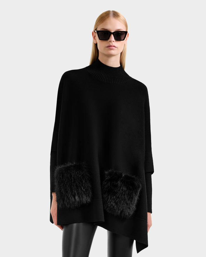 A woman in straight blonde hair models the Rudsak SWOON - 8525672 Black sweater poncho with two faux fur pockets, styled with sunglasses and black leather pants against a plain light background.