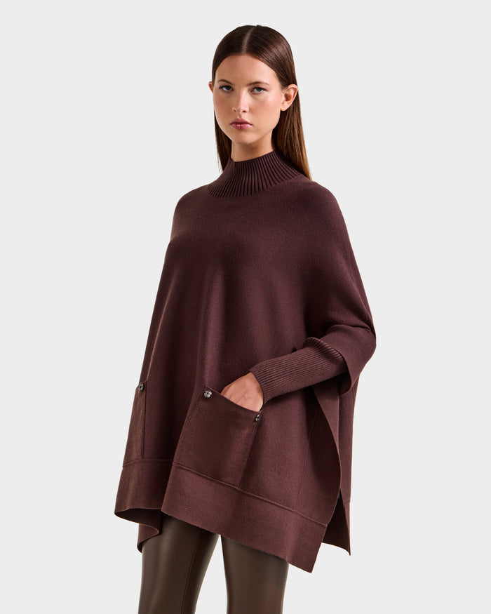 A woman wears the Rudsak SWOON - 8525672 Mocha sweater poncho with a ribbed high neckline, front pockets, and long sleeves, paired with dark brown pants, against a plain light gray background.