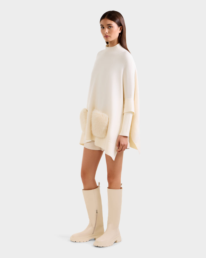 A woman models the Rudsak SWOON - 8525672 Vanilla: an oversized cream sweater poncho with faux fur pockets, paired with matching cream shorts and knee-high boots, styled against a plain light background.