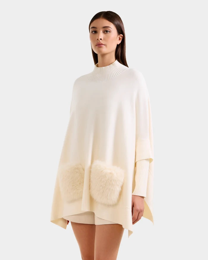 A woman models the Rudsak SWOON - 8525672 Vanilla sweater poncho, featuring a ribbed high neckline and faux fur pockets, paired with matching shorts, highlighting modern layering against a plain backdrop.