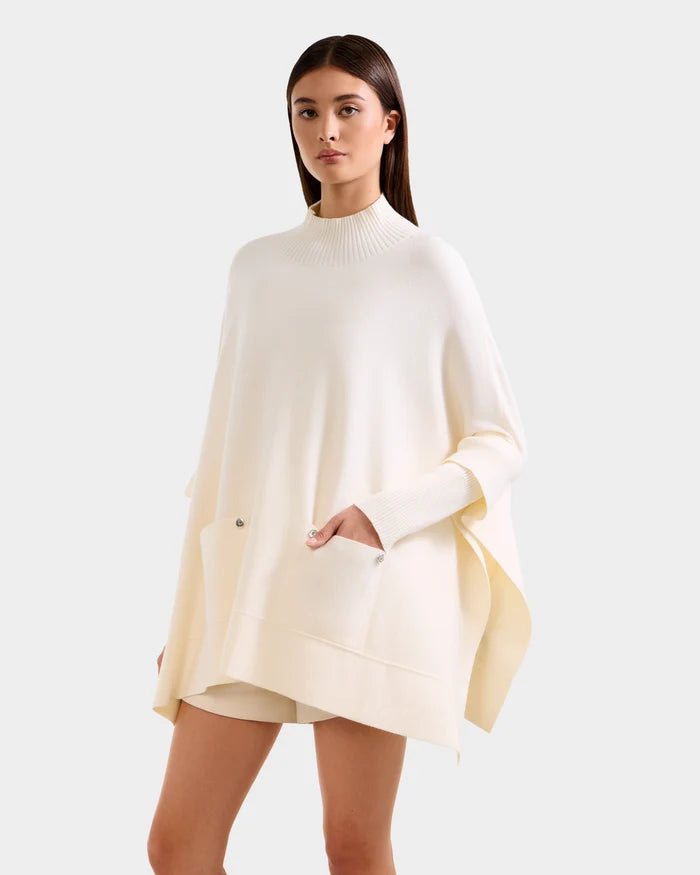 A woman stands facing forward against a light gray background, showcasing the SWOON - 8525672 Vanilla sweater poncho by Rudsak—a loose, cream-colored piece with a ribbed mock neck and front pockets for effortless modern layering.