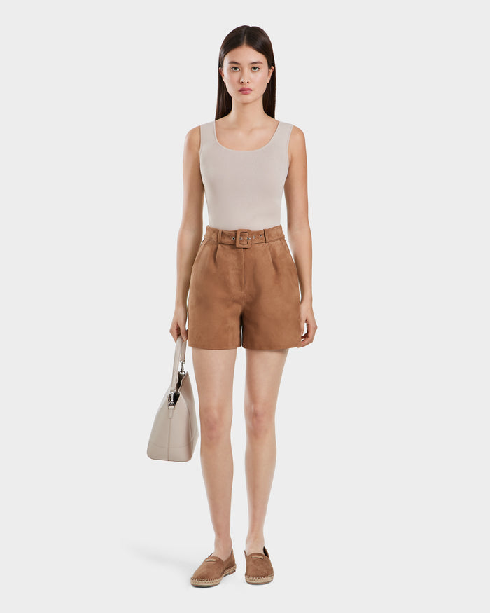 A woman with straight brown hair and a neutral expression stands facing forward, holding a light beige Rudsak TERRIE SU - 8525053 Almond handbag. She wears a sleeveless beige top, belted suede shorts, and tan espadrilles.