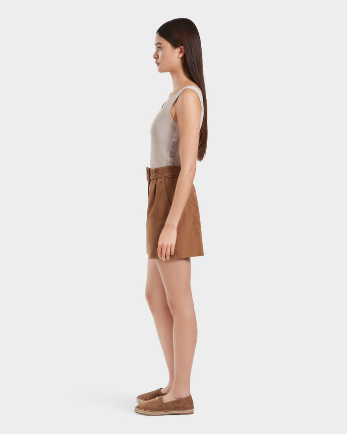 A woman with long brown hair stands in profile against a white background, wearing the Rudsak TERRIE SU - 8525053 Almond sleeveless beige top and tailored suede shorts with a belt, paired with brown espadrille shoes.