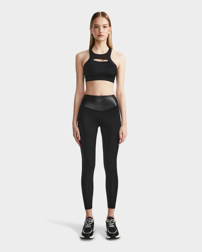 A woman faces forward in a black sports bra, Rudsak THEA - 8523003 BLACK high-rise leggings, and black athletic shoes, set against a plain light background.