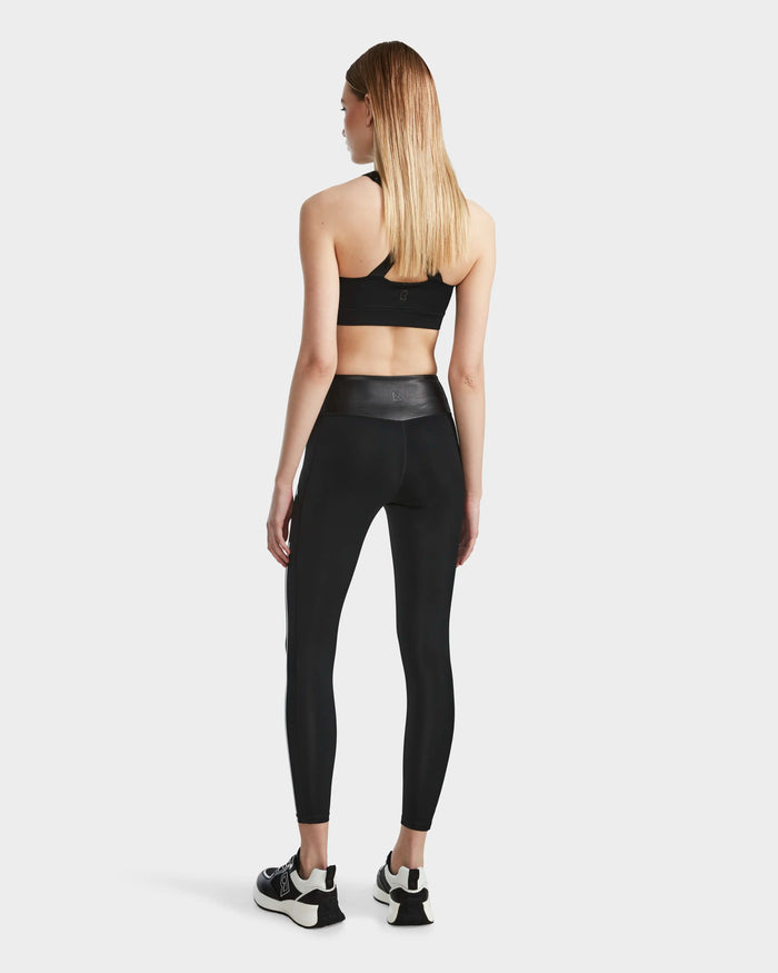 A woman with straight, shoulder-length blonde hair stands facing away in a black sports bra, black-and-white sneakers, and Rudsak THEA - 8523003 BLACK high-rise leggings against a plain white background.