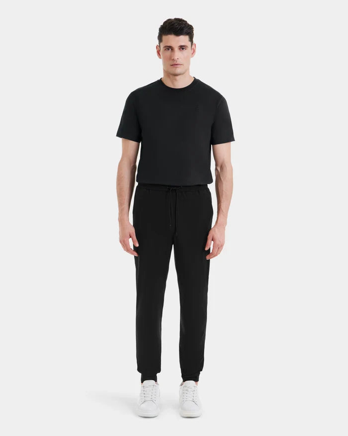 A man stands facing forward against a plain light gray background, wearing Rudsak’s TUSTIN - 6525029 Black short-sleeve t-shirt and black technical jogger pants, paired with white sneakers for an effortless men’s casual look.