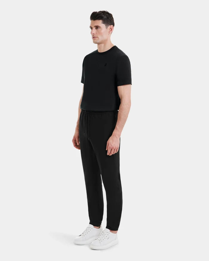 A man stands slightly to the side, wearing Rudsak’s TUSTIN - 6525029 Black jogger pants and a black T-shirt—ideal mens casualwear—with white sneakers against a plain white background.