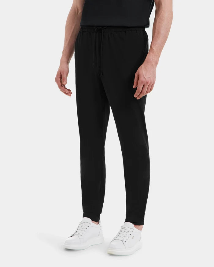 A person, shown from the shoulders down, is wearing Rudsak TUSTIN - 6525029 Black tailored jogger pants with a black shirt and white sneakers, standing against a plain white background.