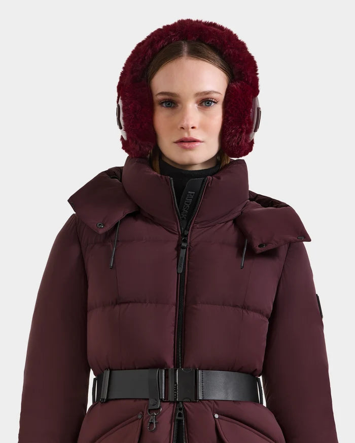 A woman stands facing forward in the Rudsak VALENCIA - 8425543 Bordeaux puffer coat with a high collar and black belt, paired with matching Rudsak faux fur earmuffs, creating a stylish winter look against a white background.