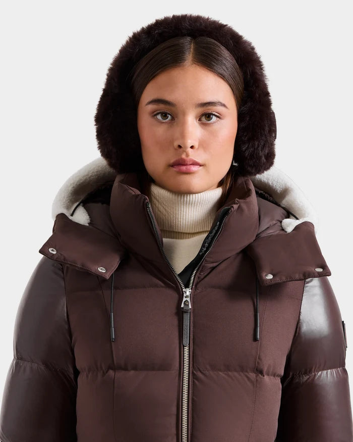 A woman with straight brown hair wears a brown puffer jacket, cream turtleneck, and Rudsak VALENCIA - 8425543 Espresso earmuffs, looking at the camera against a plain light background.