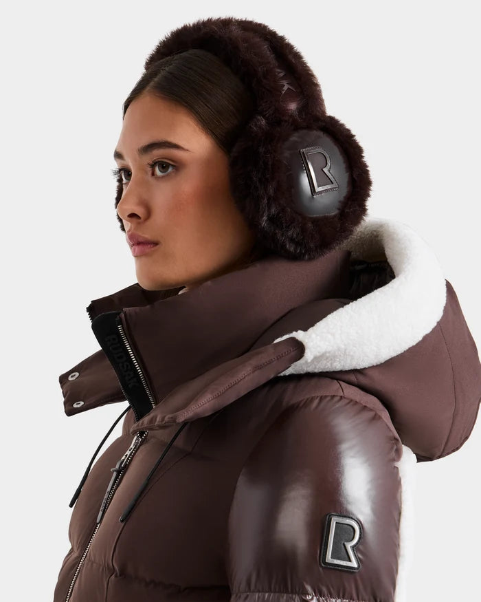 A woman in Rudsak’s VALENCIA - 8425543 Espresso earmuffs and matching brown puffer jacket with a white sherpa hood, both featuring the R logo, looks off to the side. This cozy accessory brings warmth and style against a plain background.