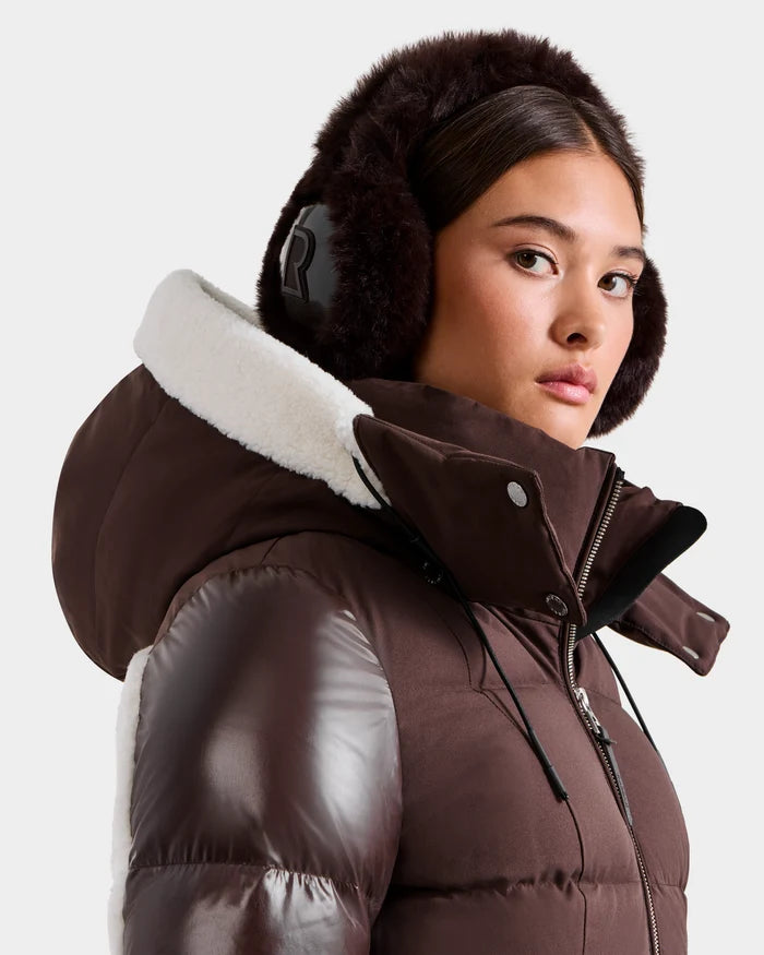 A woman wears the Rudsak VALENCIA - 8425543 Espresso brown puffer jacket with a white fleece-lined hood and large, furry earmuffs, looking to the side against a plain background to showcase this cozy winter accessory.