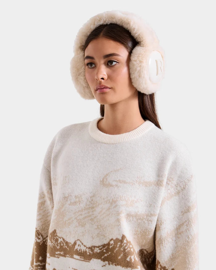 A woman with long brown hair wears Rudsak's VALENCIA - 8425543 Vanilla earmuffs and a cream sweater with a tan mountain pattern. She faces forward against a plain light gray background, highlighting this chic winter accessory.